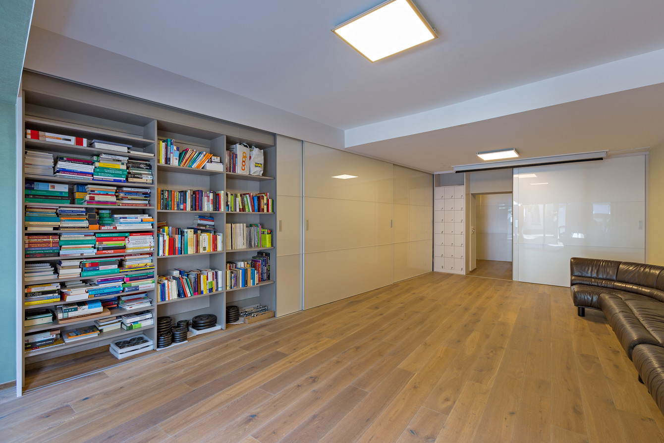 Bücherregal im Seitensystem über die ganze Wandbreite und -höhe - verschliessbar mit überbreiten Schiebetüren in Acrylglas Hochglanz 