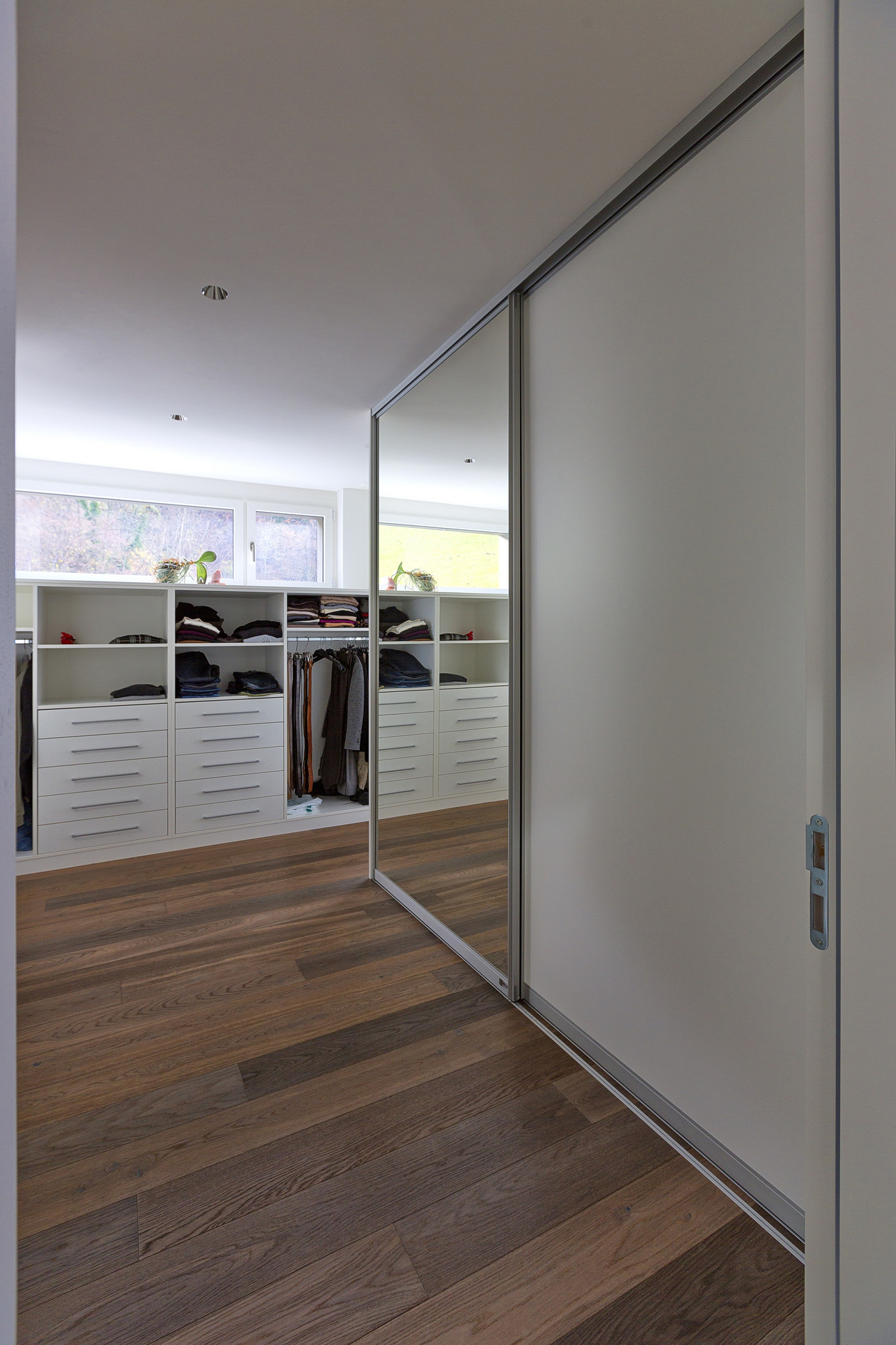 Schiebetürschrank mit Spiegel und weisser Türe als Durchgang in die offene Ankleide in weiss gehalten mit wunderschönem Holzboden in Nussholz