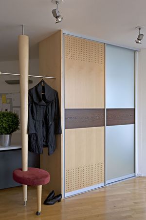 Zweitüriger Garderobenschrank mit Materialwechsel neben konischer Säule als Gästegarderobe mit drehbarem gepolsterten Sitz 